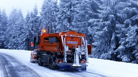 Streufahrzeug auf verschneiter Straße im Hintergrund verschneite Tannen