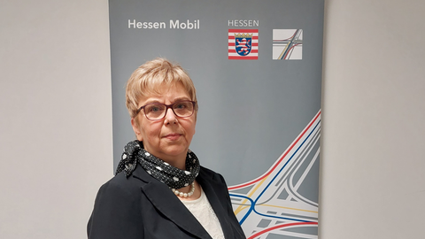 Cornelia Höhl - Regionalbüro Mittelhessen Cornelia Höhl vom Regionalbüro Mittelhessen blickt in die Kamera. Hinter Ihr ist das Logo von Hessen Mobil zu sehen.