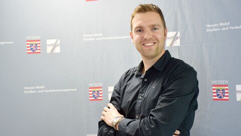Pressesprecher Nico Beck Pressesprecher Nico Beck der Region Osthessen blickt lächelend in die Kamera. Er steht vor einer grauen Logowand von Hessen Mobil.
