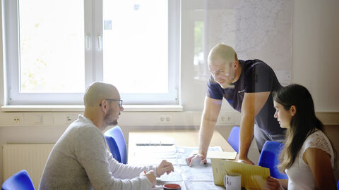 Drei Mitarbeiter von Hessen Mobil sitzen an einem Arbeitstisch und halten eine Bespechung ab. Vor Ihnen liegt ein Plan auf dem Tisch. 