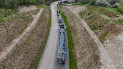 Ein Drohnenbild zeigt zwei LKW die sehr lange Brückenfertigteile aus Stahl zur Baustelle bringen. 
