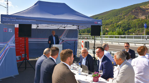Das Publikum der Verkehrsfreigabe sitzt bei herrlichem Sonnenschein an Festbankgarnituren