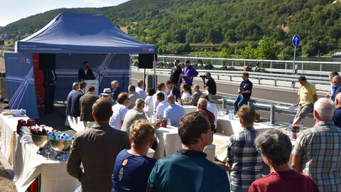Das Publikum der Verkehrsfreigabe sitzt bei herrlichem Sonnenschein an Festbankgarnituren