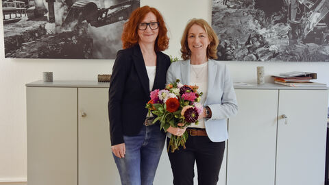 Die neue Leiterin der Abteilung Z (Zentrale Dienste) - Sylvia Hipfl-Träger  Zwei Frauen vor einem Wandbild. Die eine überreicht der anderen einen bunten Blumenstrauß