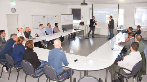 Große Runde in einem Besoprechungssall - 2 Personen stehen vorne und begrüßen die Anwesenden