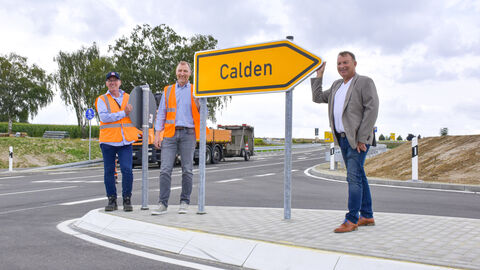 3 Personen stehen auf einer Verkehgrsinsel vor einem gelben Richtungsanzeigen auf dem Calden steht